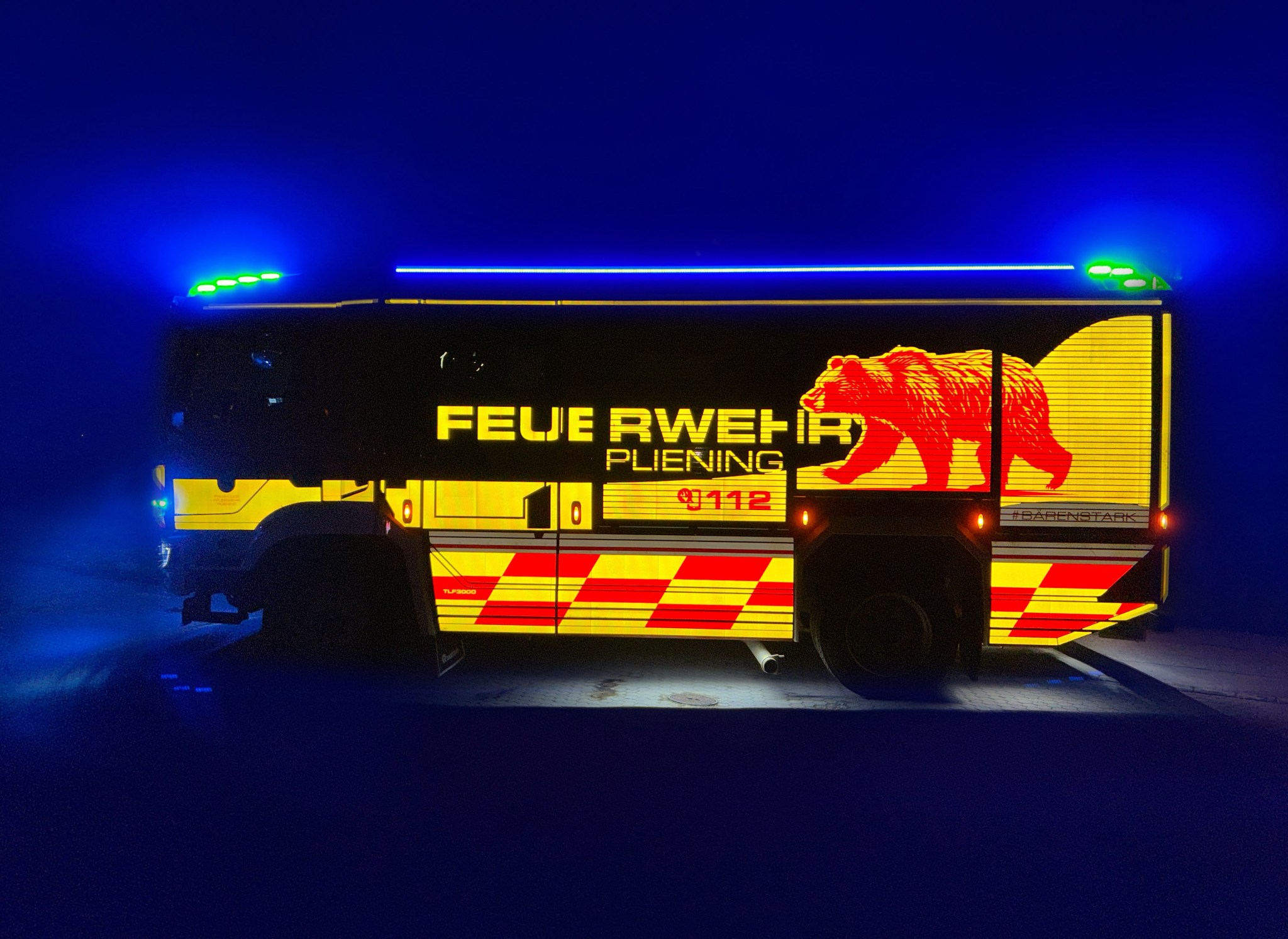 Das neue Löschfahrzeug der Feuerwehr Pliening bei Nacht von der Seite, mit leuchtender Beschriftung, rotem Bären-Motiv und rot-gelber Warnmarkierung